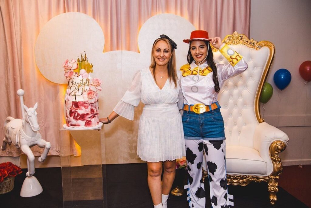 Two women posing by a Mickey Mouse backdrop with a birthday cake.