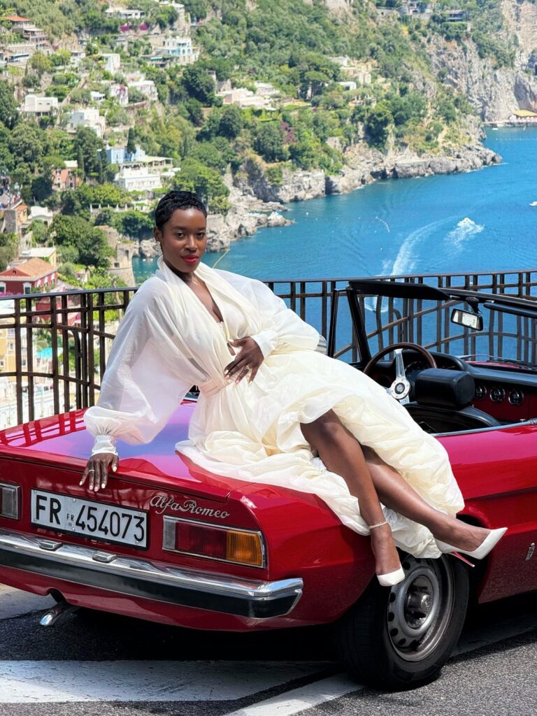Woman in white dress posing on a red vintage car overlooking Amalfi Coast