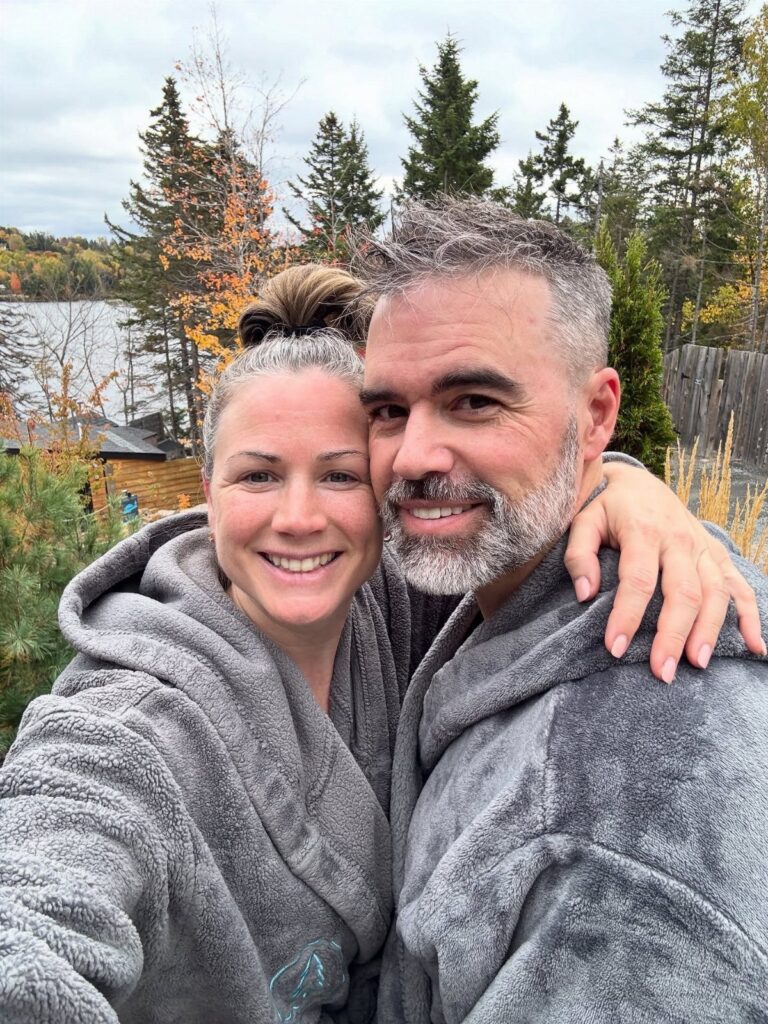 Smiling middle-aged couple in grey bathrobes posing outdoors by a lake and forest.