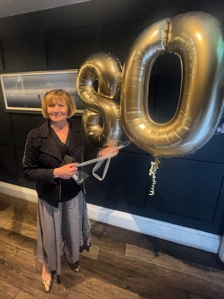 Older woman holding large gold foil balloons shaped like the number 80.