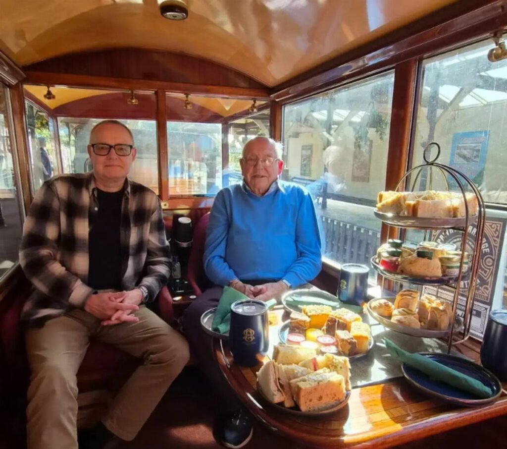 Two older men enjoying afternoon tea inside a vintage wooden train carriage.