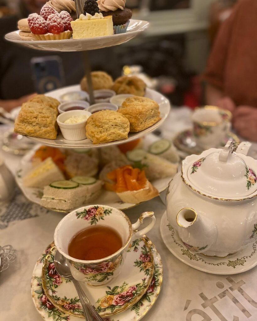 Three-tiered stand with cakes, scones, and sandwiches next to a floral teacup and teapot.