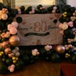 Rose gold and black balloon arch surrounding an 80th birthday banner indoors.
