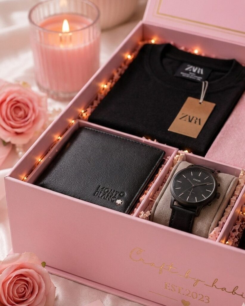 Pink gift box with black t-shirt, wallet, and watch surrounded by roses.