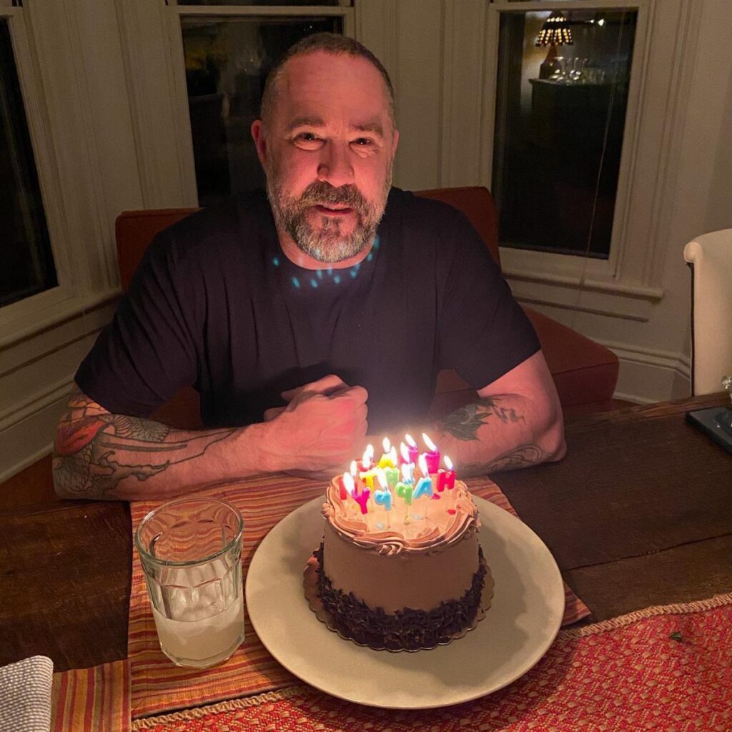 Middle-aged man with tattoos sitting behind a chocolate birthday cake with lit candles.