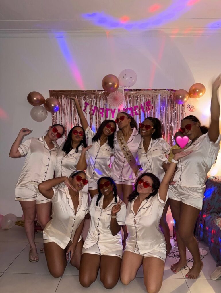 Women in white satin pajamas and heart sunglasses posing before a birthday backdrop.