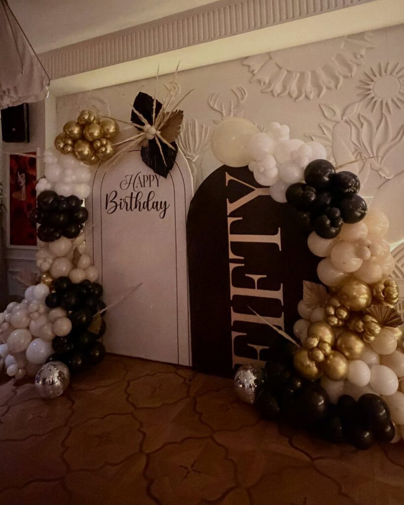 Birthday decoration with black, white, and gold balloons and 'Happy Birthday Fifty' arches.