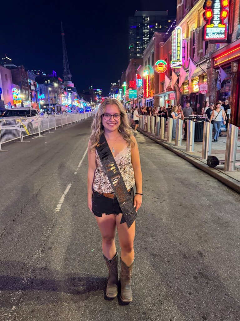 Young woman in cowboy boots and sash standing on a neon-lit Nashville street.