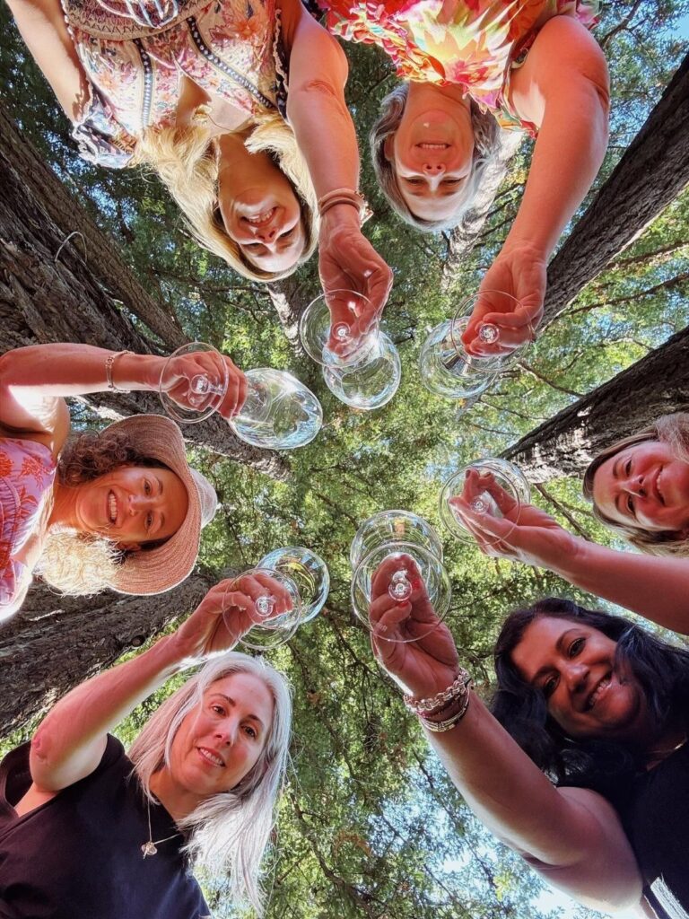 Five women clinking wine glasses in a circle under tall forest trees.
